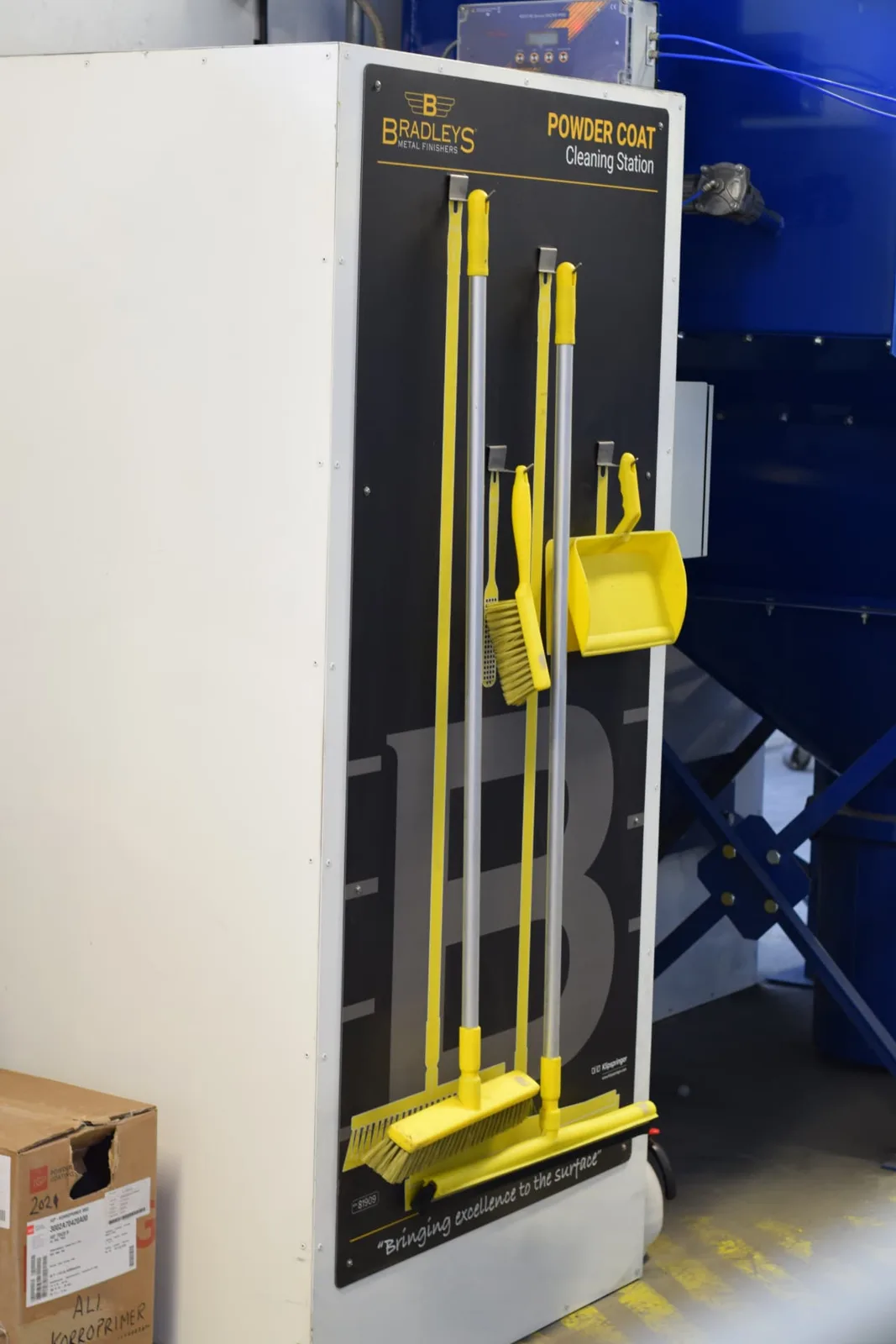 Powder coat cleaning station with yellow brushes, dustpan and squeegee mounted on a machine enclosure